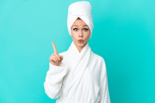 Teenager blonde girl in a bathrobe over isolated blue background intending to realizes the solution while lifting a finger up