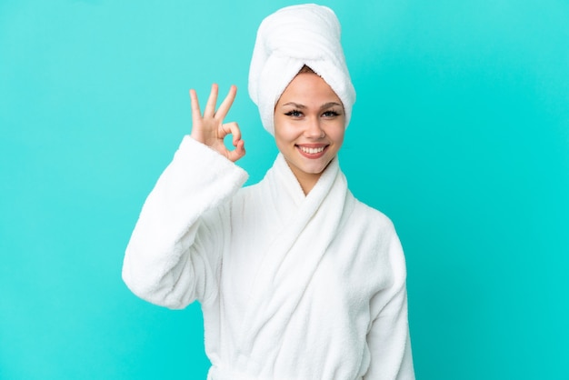 Teenager blonde girl in a bathrobe over isolated blue background showing ok sign with fingers