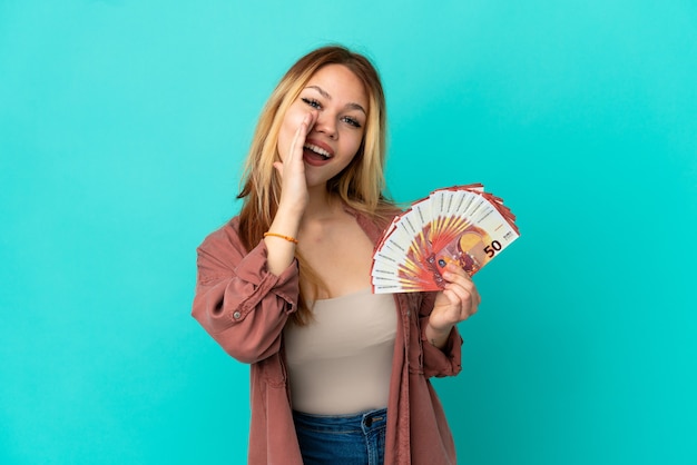 Teenager blonde girl taking a lot of Euros over isolated blue background shouting with mouth wide open