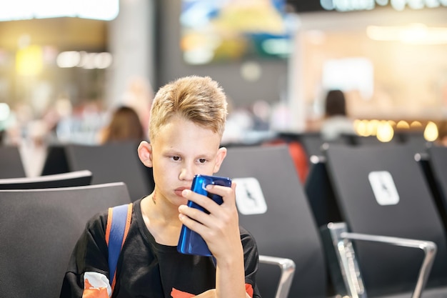 Teenager boy with backpack frowns with serious face expression and reads ebook via phone in airport