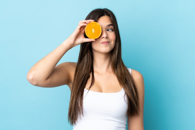 Teenager Brazilian girl over isolated blue background holding an orange