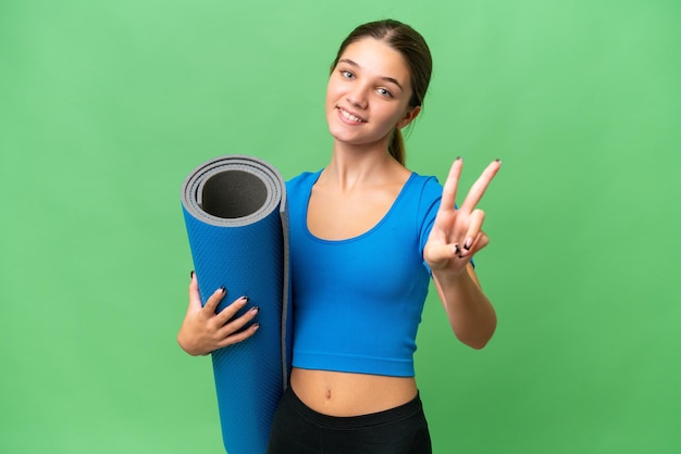 Teenager caucasian girl going to yoga classes while holding a mat over isolated background smiling and showing victory sign