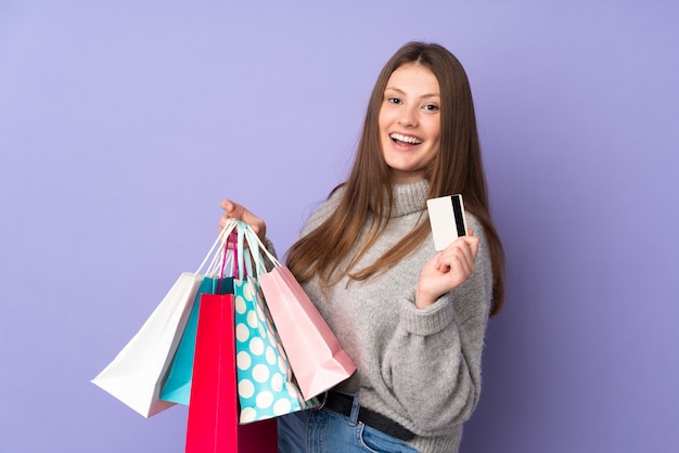 Teenager caucasian girl isolated on purple wall holding shopping bags and a credit card