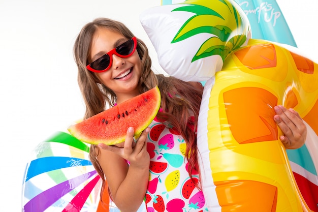 Teenager caucasian girl in sunglasses with watermelon and rubber mattresses on white wall