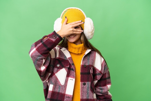 Teenager caucasian girl wearing winter muffs over isolated background covering eyes by hands and smiling