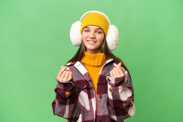 Teenager caucasian girl wearing winter muffs over isolated background making money gesture