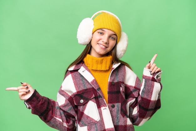 Teenager caucasian girl wearing winter muffs over isolated background pointing finger to the laterals and happy