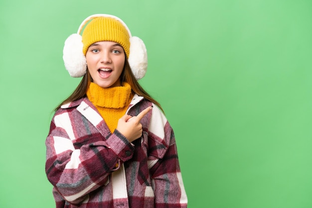 Photo teenager caucasian girl wearing winter muffs over isolated background surprised and pointing side