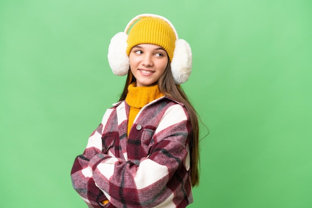 Teenager caucasian girl wearing winter muffs over isolated background with arms crossed and happy