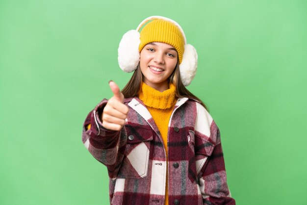 Teenager caucasian girl wearing winter muffs over isolated background with thumbs up because something good has happened