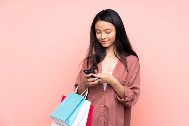 Teenager Chinese girl isolated on pink holding shopping bags and writing a message with her cell phone to a friend