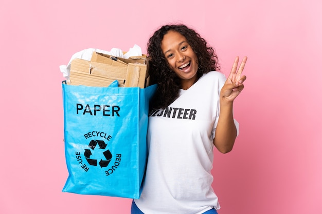 Teenager cuban girl holding a recycling bag full of paper to recycle isolated on pink  smiling and showing victory sign