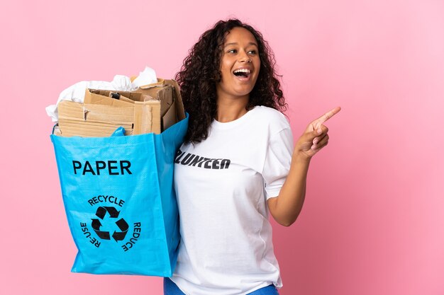 Teenager cuban girl holding a recycling bag full of paper to recycle isolated on pink wall intending to realizes the solution while lifting a finger up