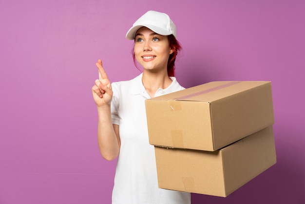 Teenager delivery girl isolated on purple background with fingers crossing and wishing the best