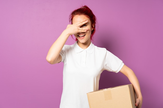 Photo teenager delivery girl on purple wall covering eyes by hands and smiling