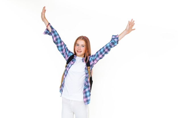 Photo teenager girl 11 years old schoolboy looking at the camera on a white background with a backpack and smiling holding up her hands high. dressed in plaid shirt. copy space