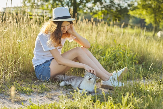 Teenager girl in hat on nature playing with gray fluffy cat, countryside