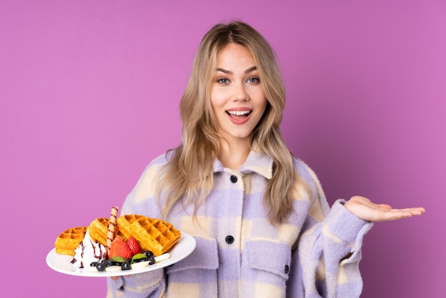 Teenager girl holding waffles on purple wall with shocked facial expression