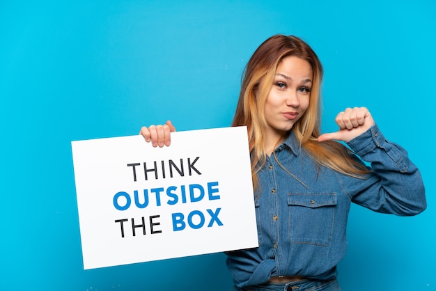 Teenager girl over isolated blue background holding a placard with text Think Outside The Box with proud gesture