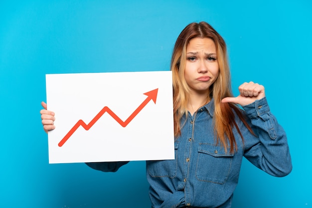 Teenager girl over isolated blue background holding a sign with a growing statistics arrow symbol with proud gesture