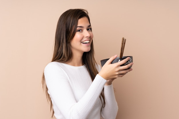 Teenager girl over isolated wall holding a bowl of noodles with chopsticks