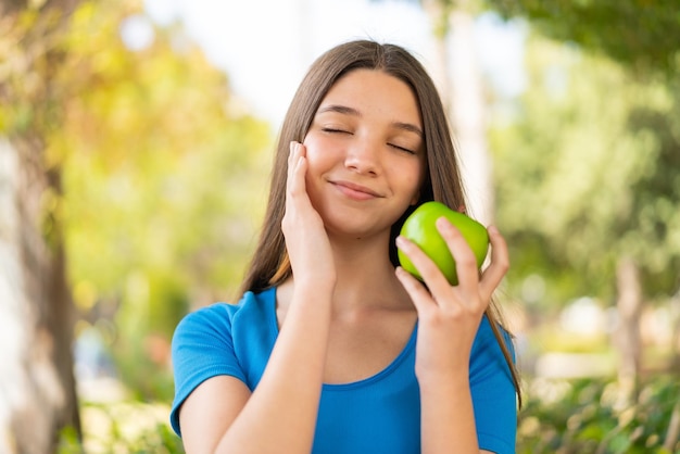 Teenager girl at outdoors holding an apple with happy expression