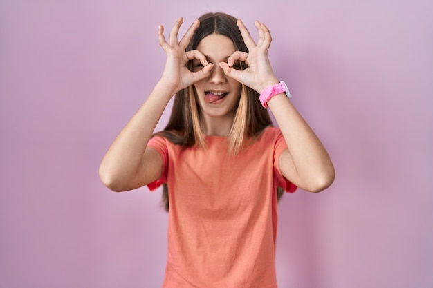 Teenager girl standing over pink background doing ok gesture like binoculars sticking tongue out eyes looking through fingers crazy expression