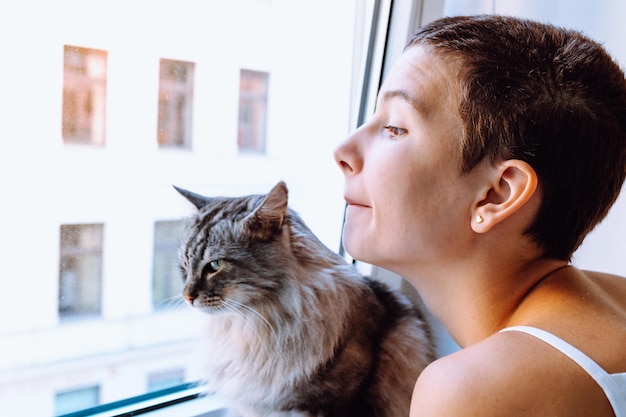 teenager girl, together with beloved domestic gray cat, look out window with curiosity