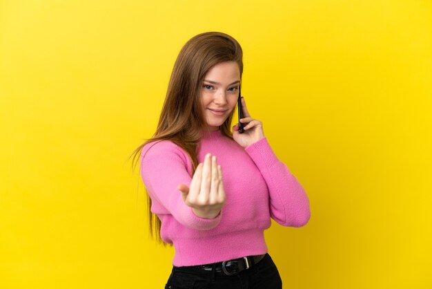 Teenager girl using mobile phone over isolated yellow background inviting to come with hand. Happy that you came