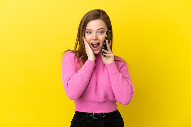 Teenager girl using mobile phone over isolated yellow background with surprise and shocked facial expression