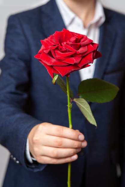 A teenager holds a red rose in his hand. The boy is dressed in a blue suit and white shirt. Close-up.