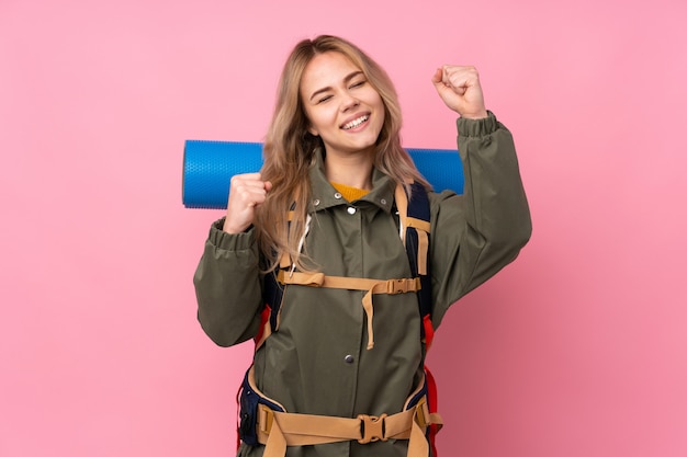 Teenager mountaineer girl with a big backpack isolated on pink celebrating a victory