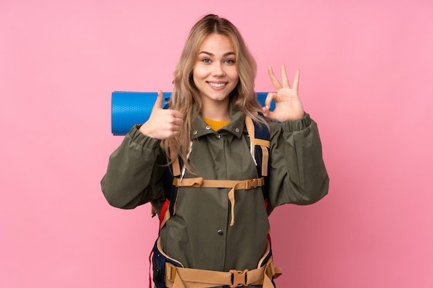 Teenager mountaineer girl with a big backpack on pink wall showing ok sign and thumb up gesture