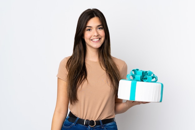 Teenager pastry chef holding a big cake over isolated white wall smiling a lot