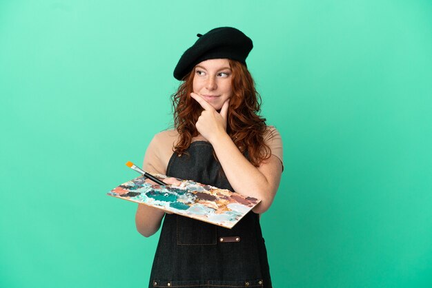 Teenager redhead artist holding a palette isolated on green background having doubts