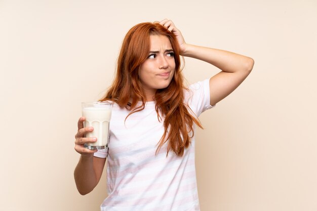 Teenager redhead girl holding a glass of milk over isolated  having doubts and with confuse face expression