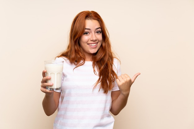 Teenager redhead girl holding a glass of milk over isolated wall pointing to the side to present a product