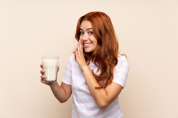 Teenager redhead girl holding a glass of milk over isolated wall whispering something