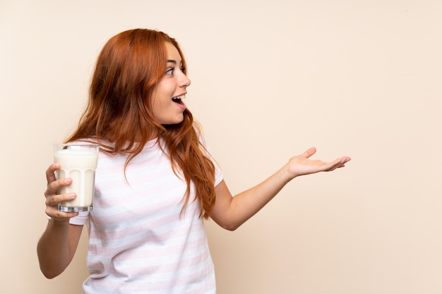 Teenager redhead girl holding a glass of milk over isolated wall with surprise facial expression