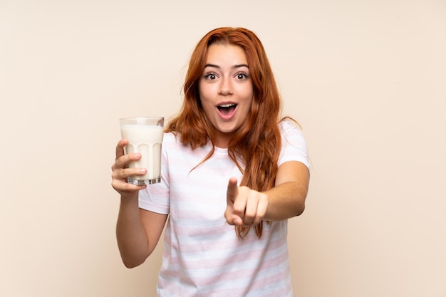 Teenager redhead girl holding a glass of milk surprised and pointing front