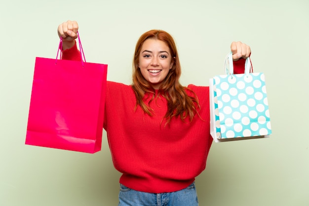 Teenager redhead girl with sweater over isolated green wall holding a lot of shopping bags