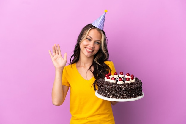 Teenager Russian girl holding birthday cake isolated on purple background saluting with hand with happy expression