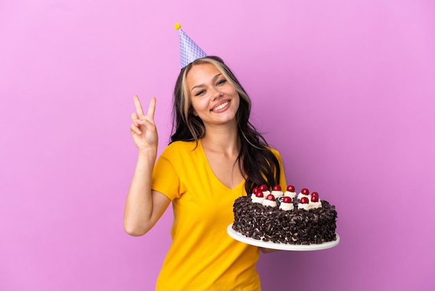 Teenager Russian girl holding birthday cake isolated on purple background smiling and showing victory sign