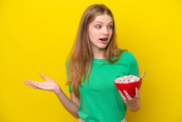 Teenager Russian girl holding bowl of cereals isolated on yellow background with surprise expression while looking side