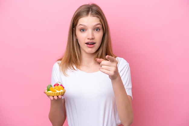 Teenager Russian girl holding a tartlet isolated on pink background surprised and pointing front