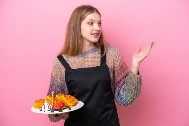 Photo teenager russian girl holding a waffles isolated on pink background extending hands to the side for inviting to come