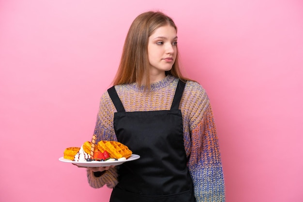 Teenager Russian girl holding a waffles isolated on pink background looking to the side