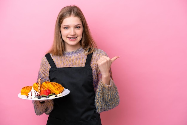 Teenager Russian girl holding a waffles isolated on pink background pointing to the side to present a product