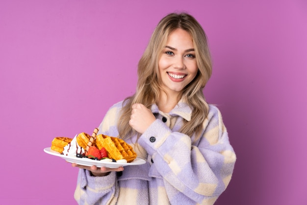 Teenager Russian girl holding waffles on purple wall celebrating a victory