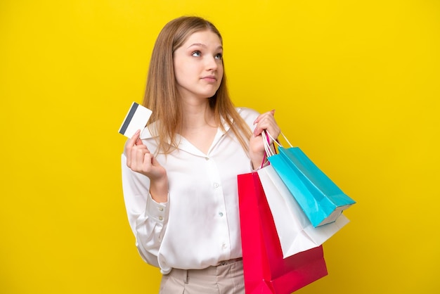 Teenager Russian girl isolated on yellow background holding shopping bags and a credit card and thinking